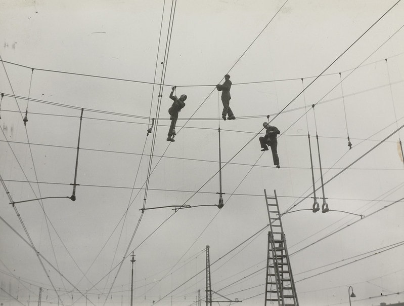 Drie mannen die op de elektriciteitslijnen van het nog in aanleg zijnde Middennet balanceren om die lijnen in orde te maken. Er staat een ladder naast waarmee ze waarschijnlijk op de elektriciteitslijnen zijn gekomen.