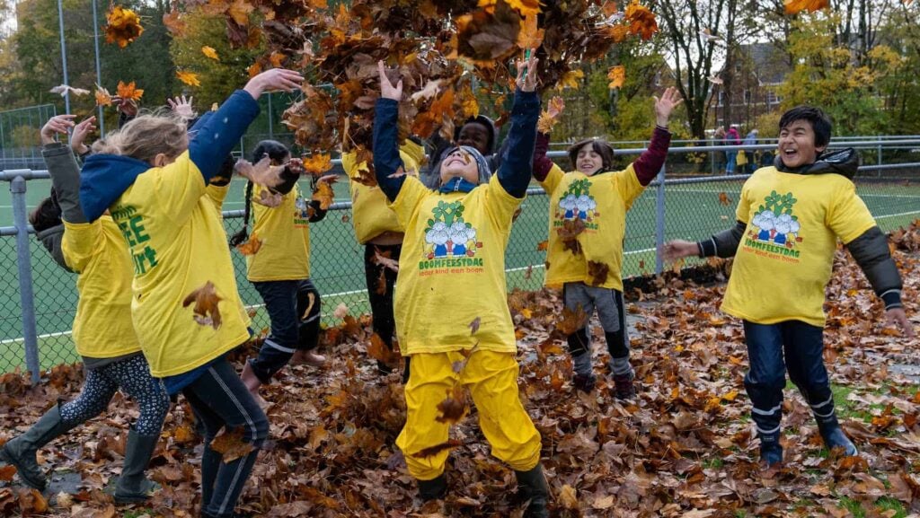 Een groepje van zeven kinderen, allemaal met een geel t-shirt aan met daarop 'boomfeestdag', staat buiten het hek van een sportveld buiten in de boombladeren. Een aantal van hen gooit een groot aantal bladeren in de lucht, en een je ziet een aantal kinderen heel vrolijk lachen.